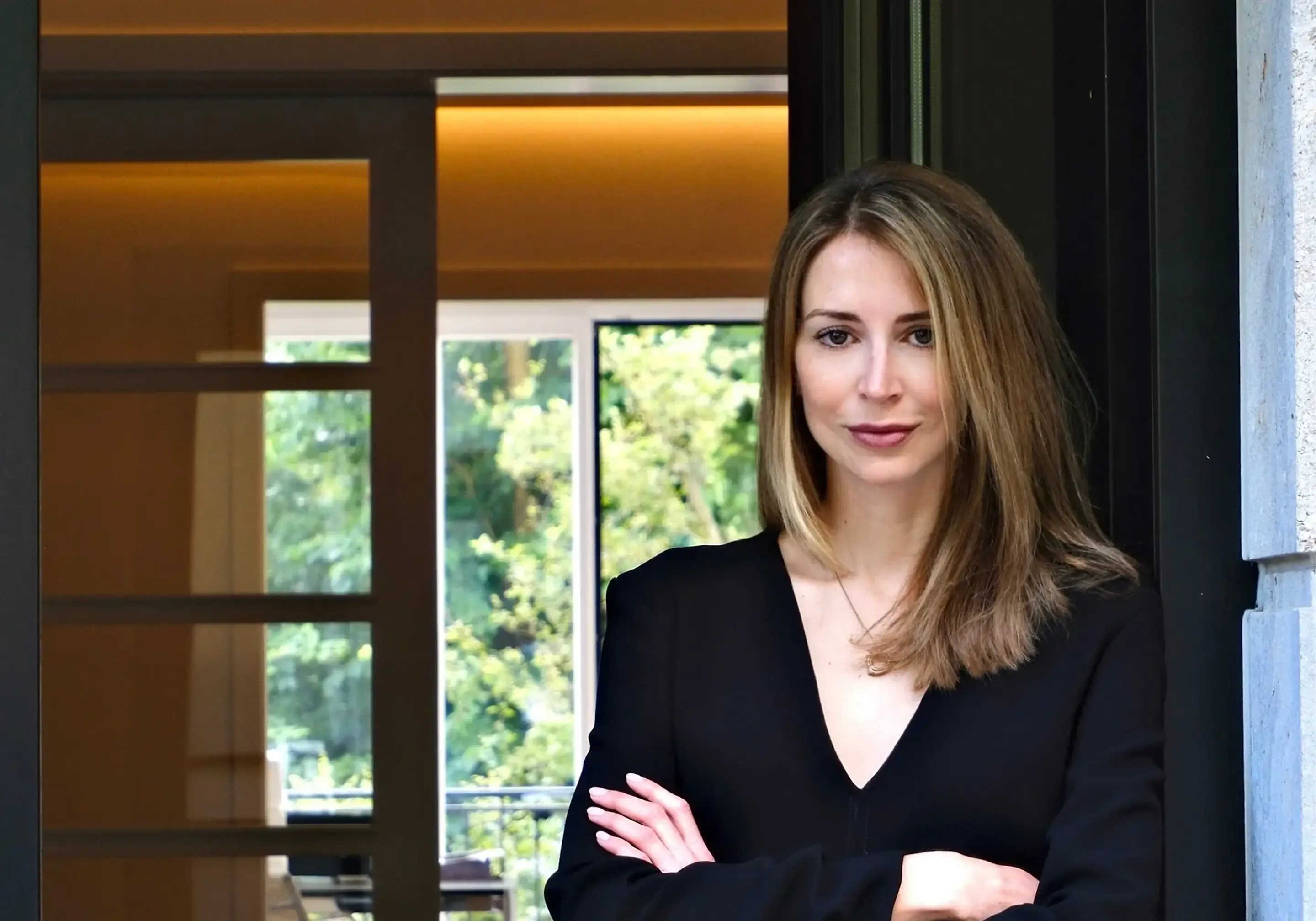 Dr. med. Lucija Grittern, Fachärztin für Dermatologie, im schwarzen Oberteil vor einem Fenster mit Blick auf grüne Bäume.