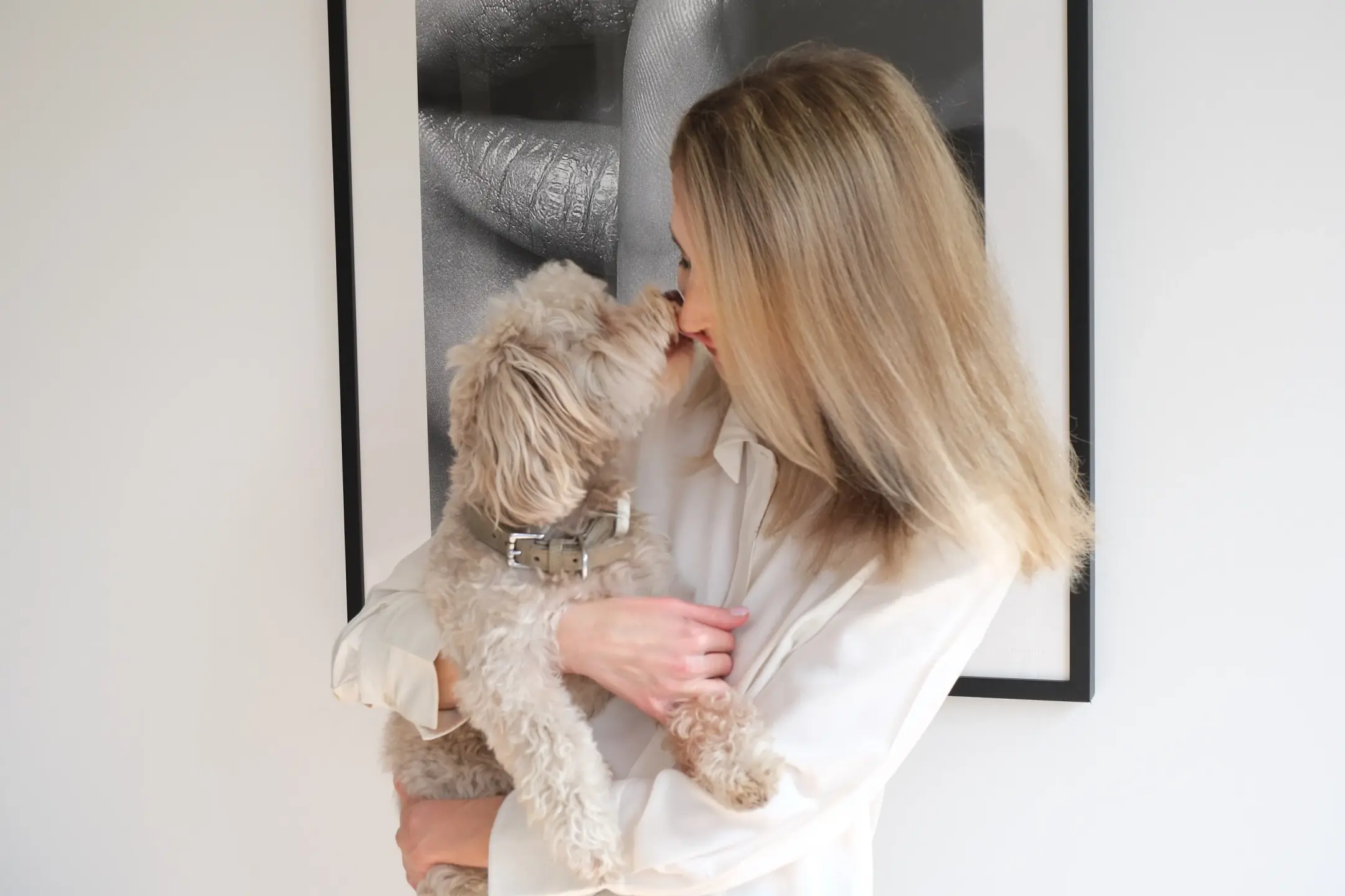 Frau in weißer Bluse hält einen kleinen Hund auf dem Arm vor einem gerahmten Bild.