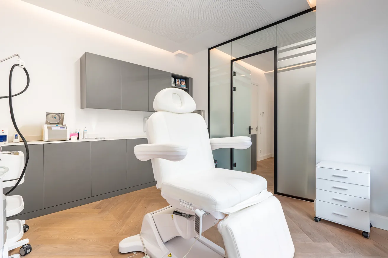 Moderne medizinische Behandlungszimmer mit verstellbarem weißen Stuhl, grauen Schränken und Glas-Eingangstür.