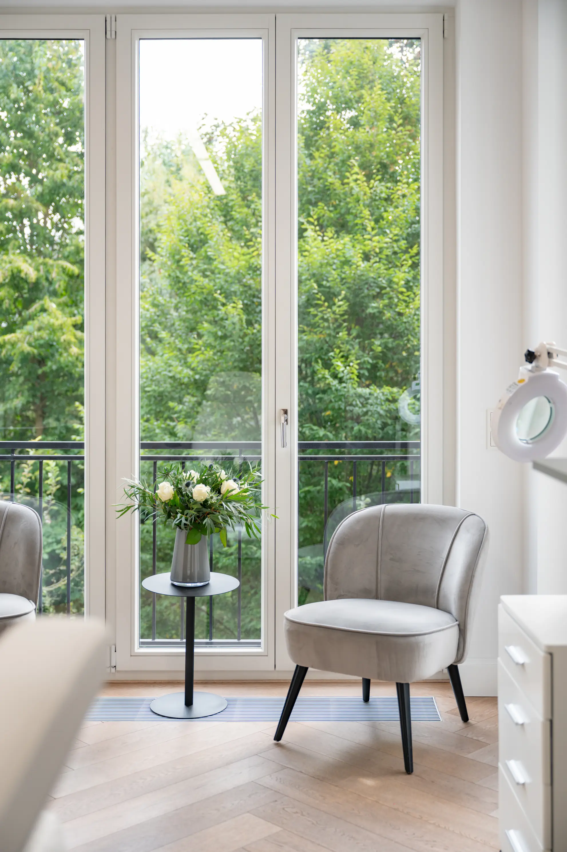 Praxisbild innen: Modernes Zimmer mit grauem Sessel, kleinem Tisch mit Blumen und großen Fenstern mit Blick auf grüne Bäume.