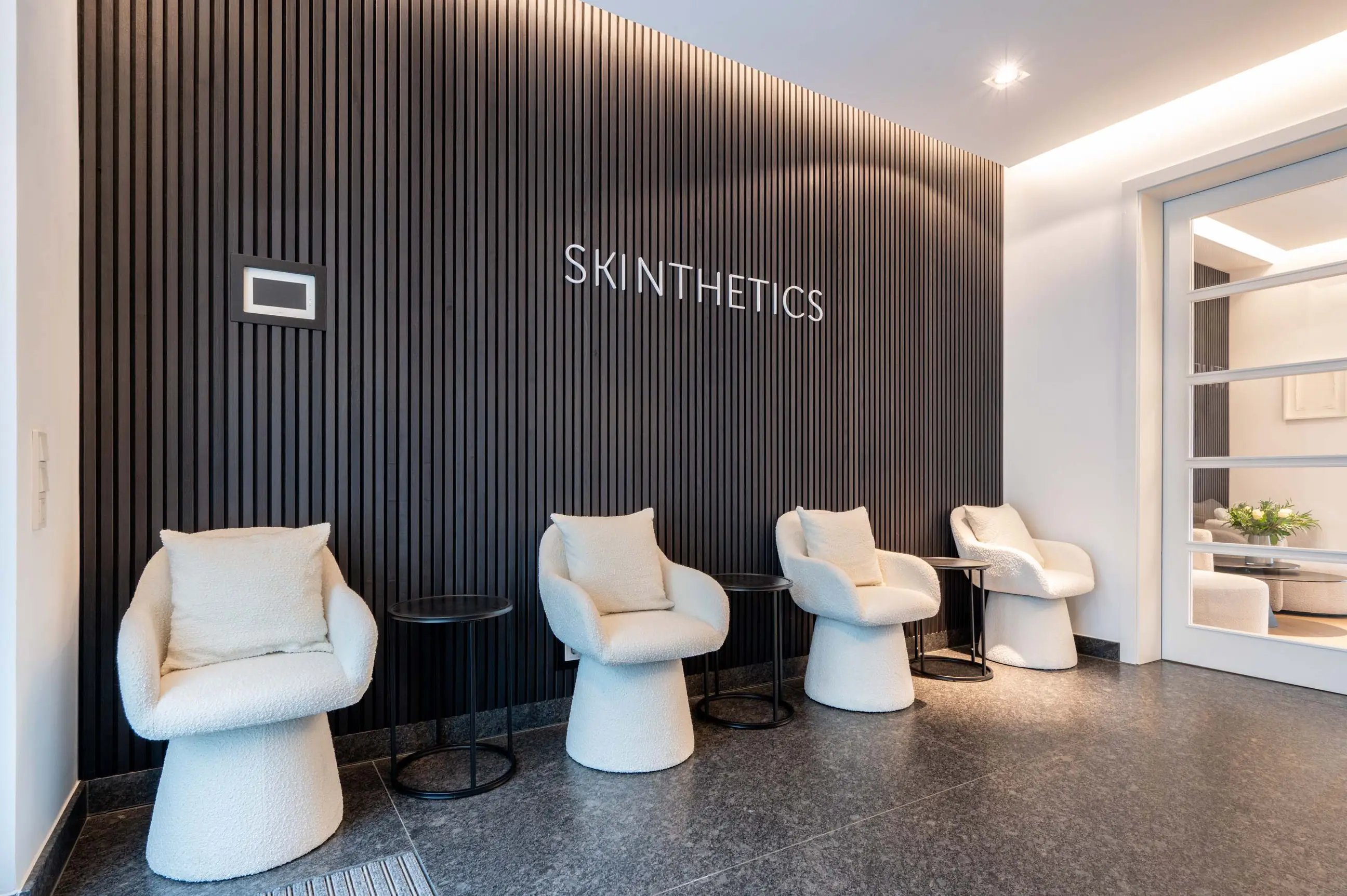 Wartebereich mit vier weißen Polstersesseln vor einer schwarzen Lamellenwand mit Schriftzug SKINTHETICS.