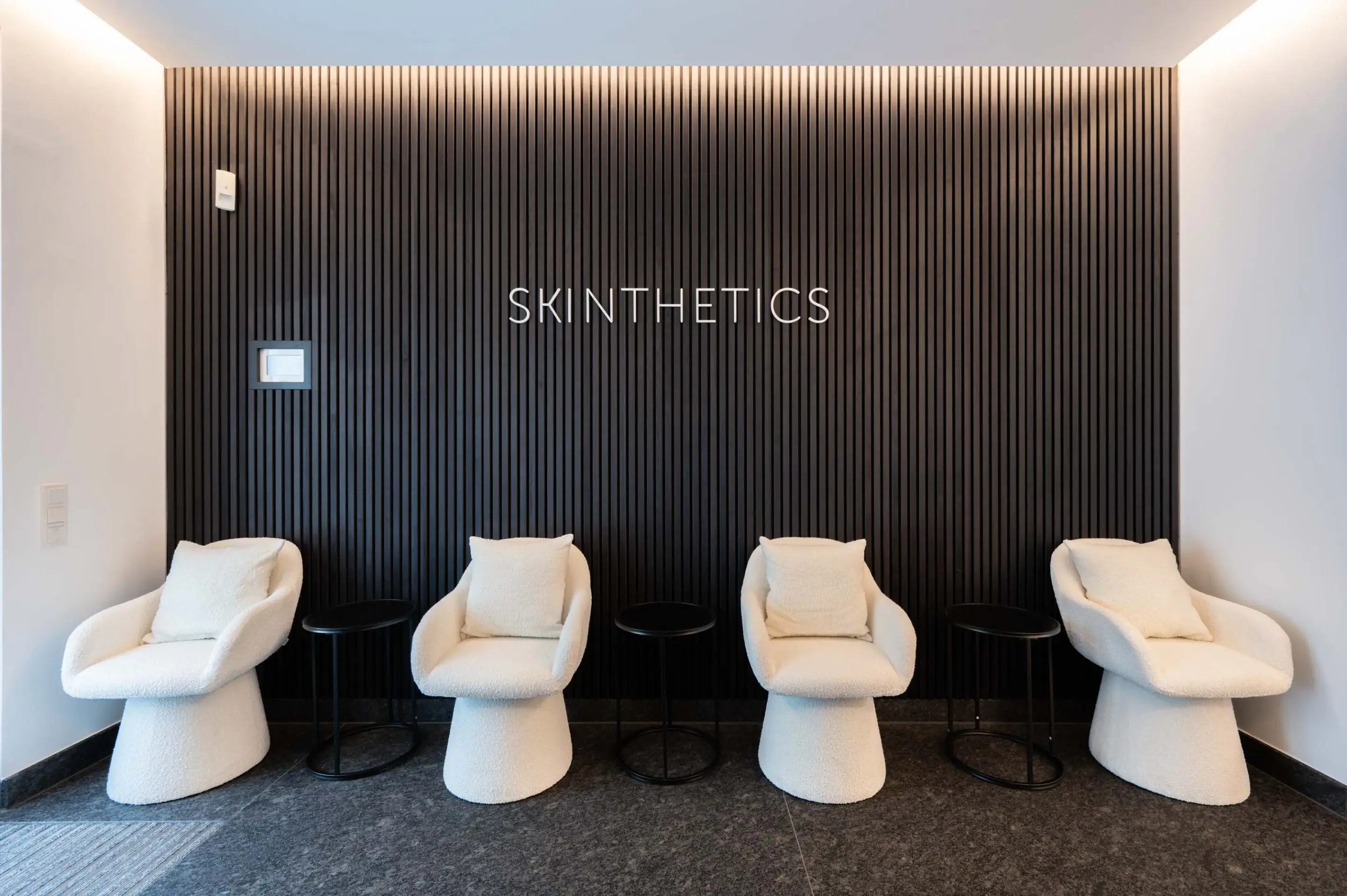 Waiting room with four white chairs with cushions in front of a black fluted wall reading SKINTHETICS.